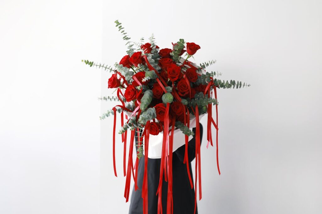 A stunning bouquet of red roses and eucalyptus, adorned with red ribbons, held by a person in formal attire.
